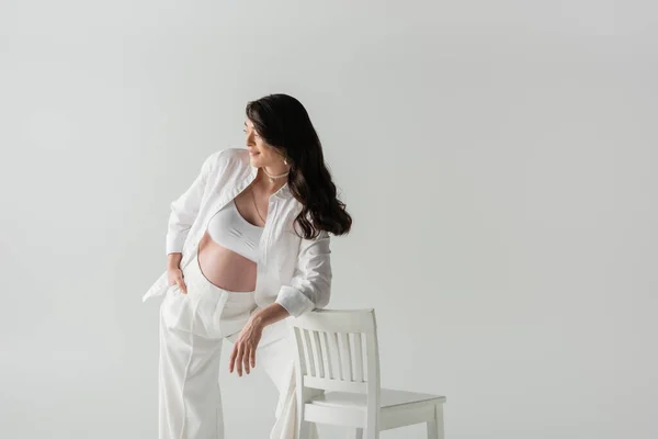 Pleased pregnant woman in fashionable pants, crop top and shirt leaning on chair and looking away isolated on grey background, maternity fashion concept — Stock Photo
