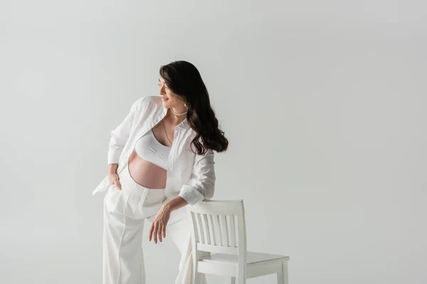 Piacere donna incinta in pantaloni alla moda, crop top e camicia appoggiata sulla sedia e guardando lontano isolato su sfondo grigio, concetto di moda maternità — Foto stock