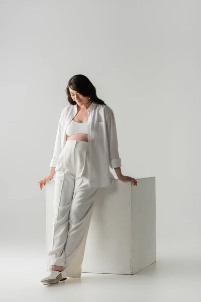 Full length of happy pregnant woman with wavy brunette hair, wearing pants, shirt and crop top, smiling near white cube on grey background, maternity fashion concept — Stock Photo