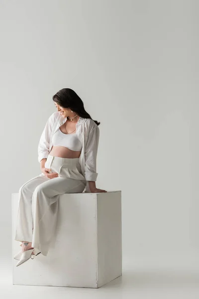 Full length of smiling mother-to-be in stylish pants, crop top and shirt touching tummy while sitting on white cube on grey background in studio, maternity fashion concept, pregnant woman — Stock Photo