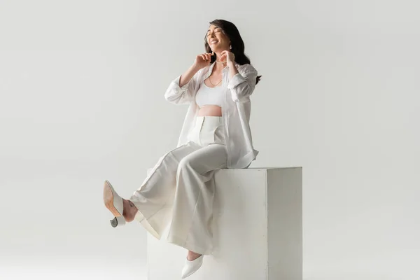 Full length of joyful expecting mother in trendy pants, shirt and cropped top sitting on white cube and smiling with closed eyes on grey background, maternity fashion concept — Stock Photo