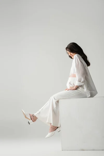 Side view of happy brunette mother-to-be in fashionable clothes such as white shirt and pants sitting on cube in studio on grey background, maternity fashion concept — Stock Photo