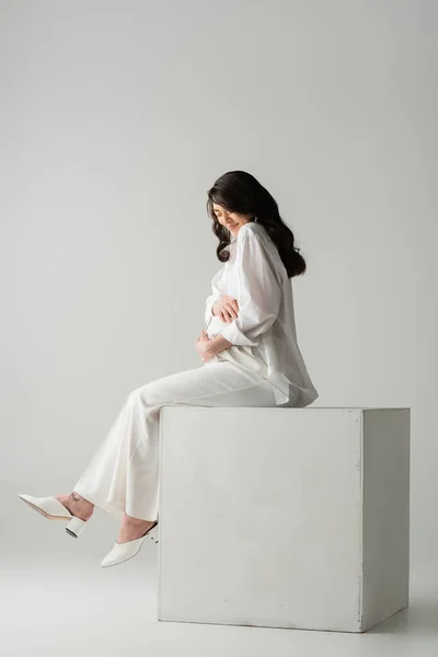 Full length of brunette pregnant woman in white shirt and pants embracing tummy while sitting on cube in studio on grey background, maternity fashion concept — Stock Photo