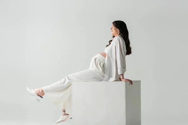Боковой вид довольной беременной женщины с волнистыми волосами брюнетки, сидящей на кубе в белых брюках, рубашке и топе на сером фоне, модная концепция материнства — стоковое фото