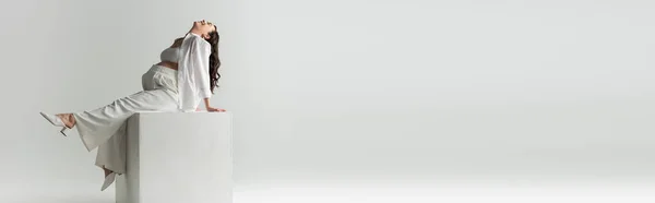 Pleine longueur de joyeuse future mère en chemise, crop top et pantalon assis sur cube blanc et souriant sur fond gris, concept de mode maternité, bannière — Photo de stock