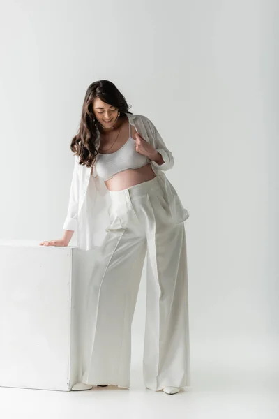 Full length of pleased pregnant woman with wavy brunette hair, wearing white pants, crop top and shirt, standing near cube on grey background, maternity fashion concept — Stock Photo