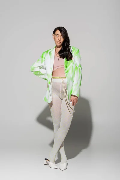 Full length of pretty brunette mother-to-be in green and white jacket, crop top, beads belt and tights posing with hand on hip and looking at camera on grey background, trendy pregnancy concept — Stock Photo