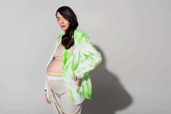 Будущая мама с волнистыми волосами брюнетки, в зеленой и белой куртке, с обрезками и леггинсами, позируя рукой на бедре на сером фоне, модная концепция беременности — стоковое фото