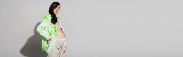 Elegante mom-to-be com cabelo morena ondulado, vestindo jaqueta verde e branco, leggings e contas cinto de pé com a mão no quadril no fundo cinza, conceito de moda maternidade, bandeira, mulher grávida — Fotografia de Stock