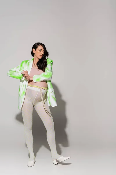 Полная длина стильная будущая мама в зеленой и белой куртке, топ, леггинсы и бусы пояса глядя в сторону на сером фоне, модная концепция беременности, беременная женщина — стоковое фото