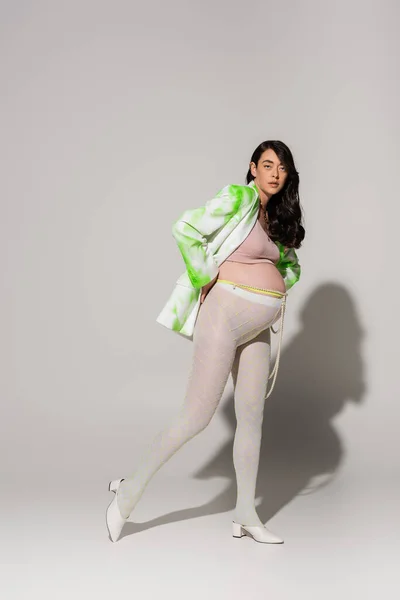 Brünette und bezaubernde Schwangere in Leggings, bauchfreiem Oberteil, grün-weißer Jacke, die auf grauem Hintergrund posiert, Umstandsmodekonzept, Erwartung — Stockfoto