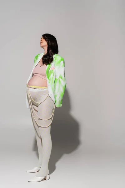 Full length of fashionable pregnant woman in tights, crop top, green and white jacket and beads belt standing on grey background, maternity fashion concept, expectation — Stock Photo