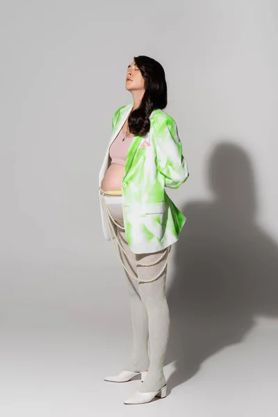 Полная длина беременной женщины с закрытыми глазами, стоящей в зелено-белой куртке, топе, поясе из бусин и леггинсах на сером фоне, концепция в стиле материнства, ожидание — стоковое фото