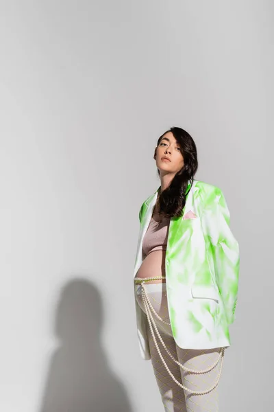 Stylische schwangere Frau mit welligem brünetten Haar, die in die Kamera schaut, während sie in grün-weißem Blazer, Crop Top, Leggings und Perlengürtel auf grauem Hintergrund posiert, Erwartung, Umstandsmodekonzept — Stockfoto