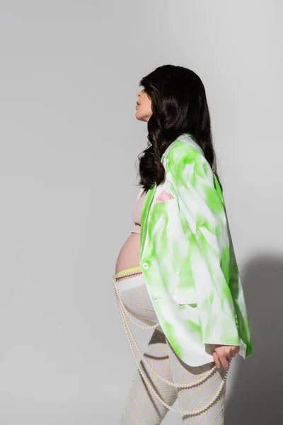 Side view of pregnant woman with wavy brunette hair posing in green and white jacket, crop top, beads belt and leggings on grey background, maternity fashion concept, expectation — Stock Photo
