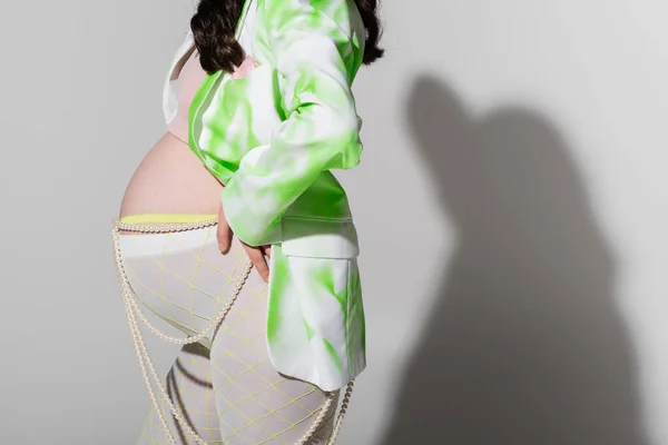 Vue recadrée de la femme enceinte en blazer vert et blanc, crop top, ceinture de perles et leggings debout avec la main sur la hanche sur fond gris, concept de maternité à la mode, attente — Photo de stock