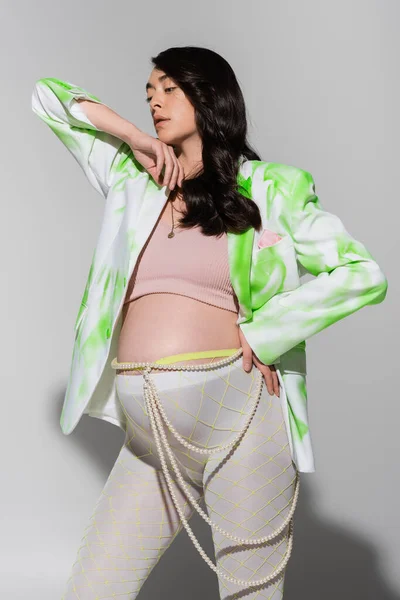 Стильная ожидающая мать в зеленой и белой куртке, топ, колготки и бусы пояса позируя с рукой на бедре на сером фоне, концепция моды материнства, беременная женщина, красивая — стоковое фото