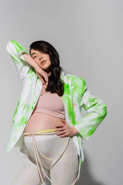 Femme enceinte avec des cheveux bruns ondulés posant dans le haut de la culture, blazer vert et blanc, ceinture de perles et leggings tout en regardant la caméra sur fond gris, concept de maternité à la mode, attente — Photo de stock