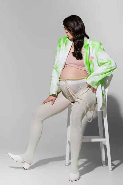 Полная длина модной будущей матери в леггинсах, поясе из бусин, топ и белый и зеленый блейзер сидя на стуле на сером фоне, концепция материнства стиль, ожидание — стоковое фото
