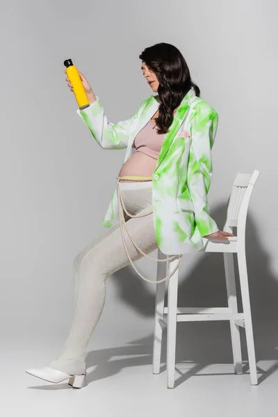 Полная длина беременной женщины с блестящими волосами брюнетки, в стильной куртке, леггинсах и поясе из бусин, сидя на стуле и держа лак для волос на сером фоне, концепция моды для беременных — стоковое фото