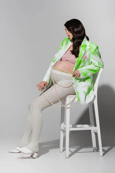 Comprimento total de morena mãe-a-ser sentado na cadeira em blazer verde e branco, top de cultura e leggings ao tocar cinto contas em fundo cinza, expectativa, conceito de moda maternidade — Fotografia de Stock