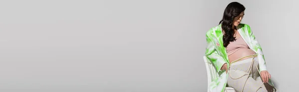Affascinante futura madre in crop top, blazer verde e bianco, perline cintura e leggings seduto sulla sedia su sfondo grigio, concetto di stile maternità, banner, aspettativa, donna incinta — Foto stock