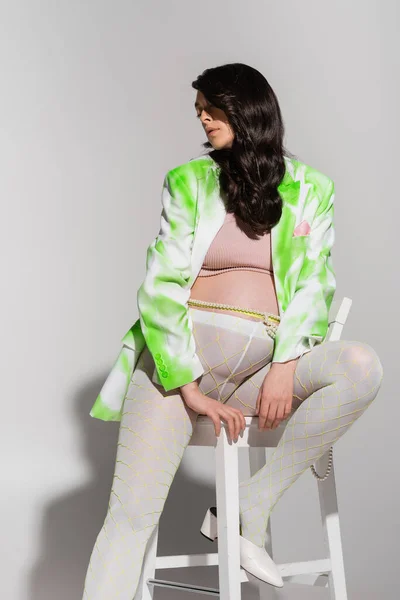 Mujer embarazada con cabello castaño ondulado, vistiendo chaqueta verde y blanca, top de la cosecha, leggins y cinturón de cuentas, posando en silla con los ojos cerrados sobre fondo gris, concepto de moda de maternidad - foto de stock