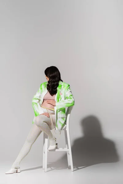 Полная длина стильная будущая мама с волнистыми брюнетками, позирующими на стуле в зеленом и белом блейзере, топ, пояс из бусин и леггинсы на сером фоне, концепция моды для беременных — стоковое фото