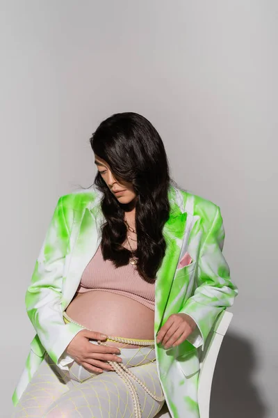 Будущая мама с волнистыми волосами брюнетки касаясь живота во время позирования в зеленом и белом блейзере, топ и бусы пояса на сером фоне, концепция моды материнства, ожидание, беременная женщина — стоковое фото