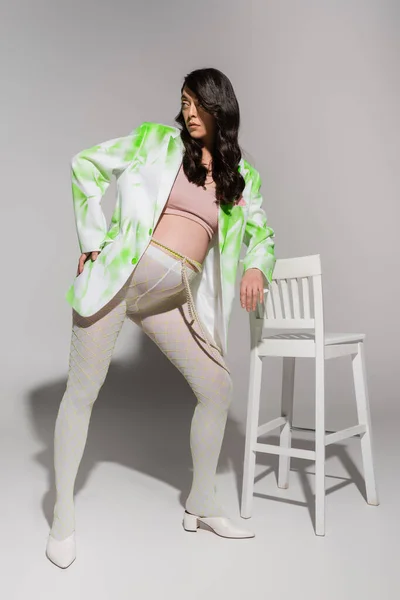 Полная длина модной беременной женщины в зеленом и белом блейзере, кукурузный топ, леггинсы и бусины пояса позируя возле стула и глядя в сторону на сером фоне, концепция материнства стиль, ожидание — стоковое фото