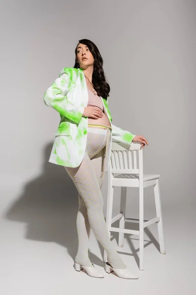 Full length of brunette pregnant woman in fashionable blazer, crop top, beads belt and leggings touching belly and looking away near chair on grey background, maternity fashion concept, expectation — Stock Photo