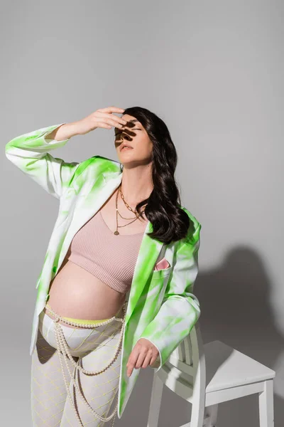 Appealing pregnant woman in crop top, green and white jacket, leggings and beads belt posing with closed eyes and hand near face on grey background, expectation, maternity fashion concept — Stock Photo