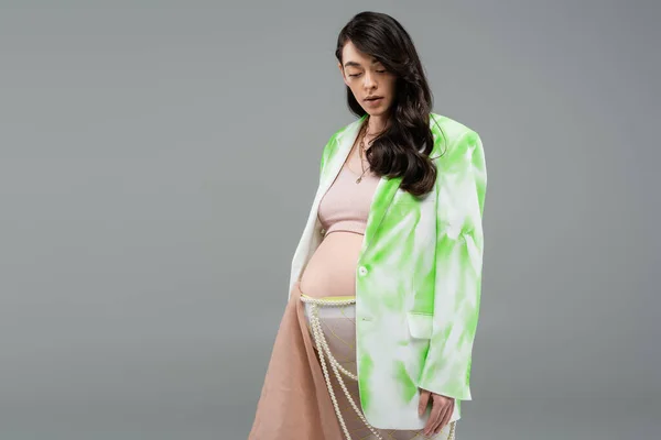 Encantadora madre embarazada en chaqueta verde y blanco, top de la cosecha y medias con tela de gasa beige y cinturón de perlas de pie aislado sobre fondo gris, concepto de maternidad de moda - foto de stock