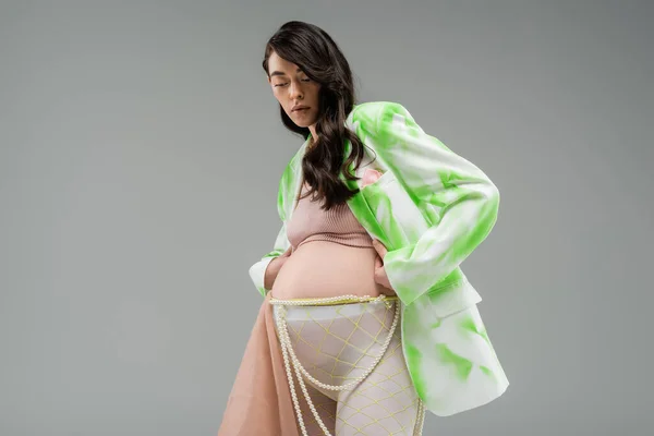 Vista a basso angolo della futura mamma bruna in giacca verde e bianca, crop top, leggings con tessuto chiffon beige e cintura di perline isolata su sfondo grigio, concetto di gravidanza alla moda, aspettativa — Foto stock