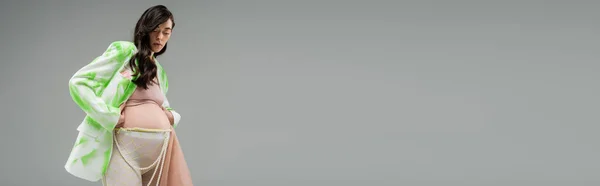 Visão de baixo ângulo da futura mãe em blazer verde e branco na moda, top de cultura, leggings com cinto de contas e pano de chiffon isolado em fundo cinza, conceito de moda gravidez, banner — Fotografia de Stock