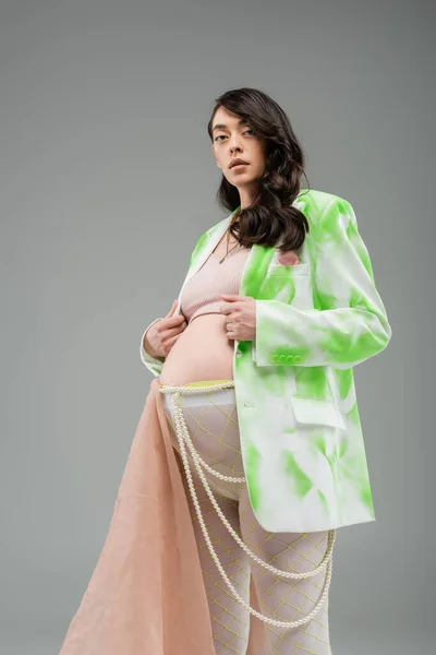 Low angle view of charming mother-to-be in green and white blazer, crop top, beads belt and leggings with chiffon cloth looking at camera isolated on grey background, fashionable maternity concept — Stock Photo