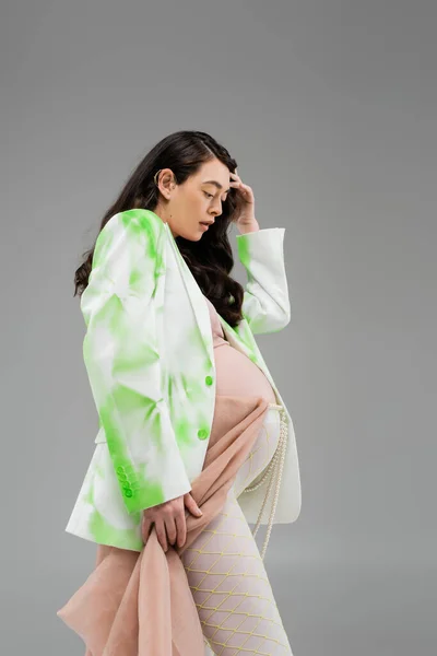 Вид збоку вагітної жінки з хвилястим брюнетка волосся позує в стильній зелено-білій куртці, легінси з поясом з бісеру і бежевий шифон тканини ізольовані на сірому фоні, концепція моди материнства — стокове фото