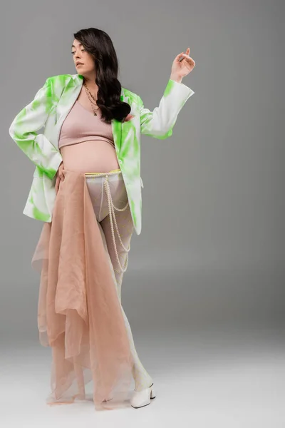 Полноразмерная брюнетка беременная женщина в зеленой и белой куртке, топ, ремень из бусин и леггинсы, позирующая с бежевой шифоновой тканью на сером фоне, концепция одежды для беременных, ожидание — стоковое фото