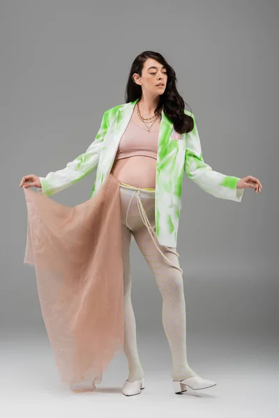 Comprimento total de morena mãe-a-ser em casaco verde e branco, top de cultura e leggings posando com pano de chiffon bege em fundo cinza, conceito de moda maternidade, expectativa — Fotografia de Stock