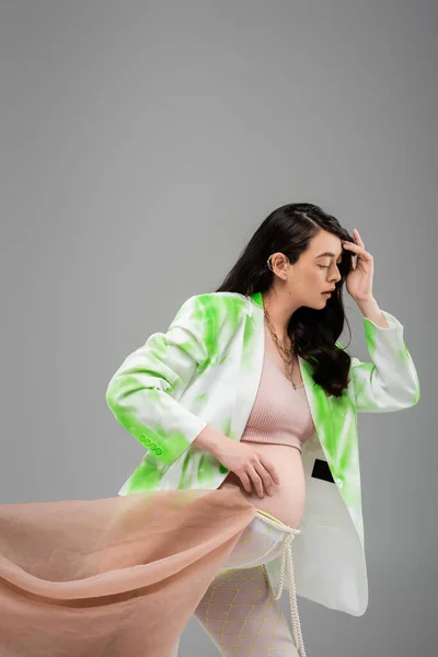 Brunette pregnant woman in green and white blazer, crop top, beads belt and leggings with chiffon cloth isolated on grey background, maternity fashion concept, expectation — Stock Photo
