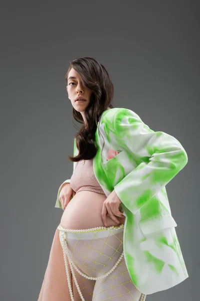 Брюнетка беременная женщина в стильной куртке, топ, ремень из бусин и леггинсы с шифоновой тканью, смотрящая на камеру, изолированную на сером фоне, концепция моды для беременных, будущая мама — стоковое фото