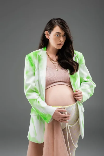 Expecting mother with wavy brunette hair, wearing green blazer, crop top and leggings with beads belt and chiffon cloth, looking at camera isolated on grey background, fashionable maternity concept — Stock Photo