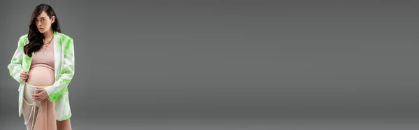 Brünette schwangere Frau im trendigen Blazer, bauchfreiem Oberteil, Perlengürtel und Leggings mit beigem Chiffontuch, Blick in die Kamera isoliert auf grauem Hintergrund, Umstandsmodekonzept, Banner — Stockfoto