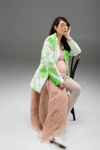 Полная длина привлекательной беременной женщины в стильной куртке, топе и леггинсах с поясом из бусин и бежевым шифоном, сидящей на стуле и смотрящей на камеру на сером фоне, концепция моды для беременных — стоковое фото