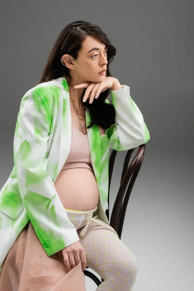 Mulher grávida morena em casaco verde e branco, top de cultura, cinto de contas e leggings sentados na cadeira e olhando para o fundo cinza, conceito de moda maternidade — Fotografia de Stock