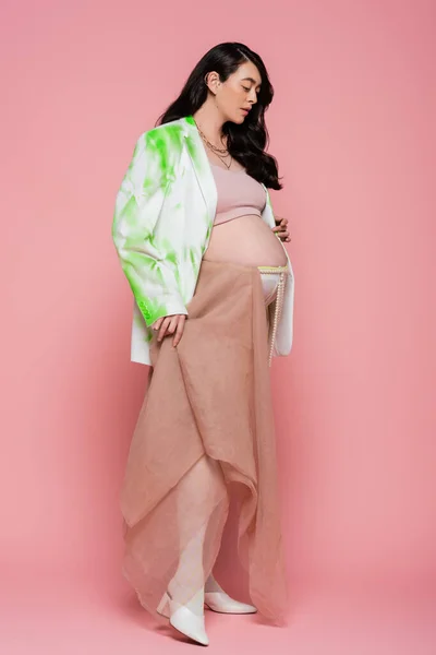 Volle Länge der werdenden Mutter in grün-weißer Jacke, bauchfreiem Oberteil, Strumpfhose mit Perlengürtel und Chiffontuch auf rosa Hintergrund, modisches Schwangerschaftskonzept, schwangere Frau — Stockfoto
