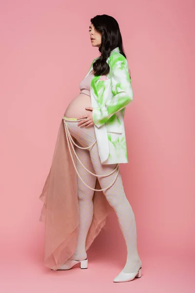 Повна довжина чарівної вагітної жінки в стильному блістері, верхній частині культури, легінси з бежевою шифоновою тканиною і поясом з бісеру, що стоїть на рожевому фоні, концепція моди материнства — стокове фото