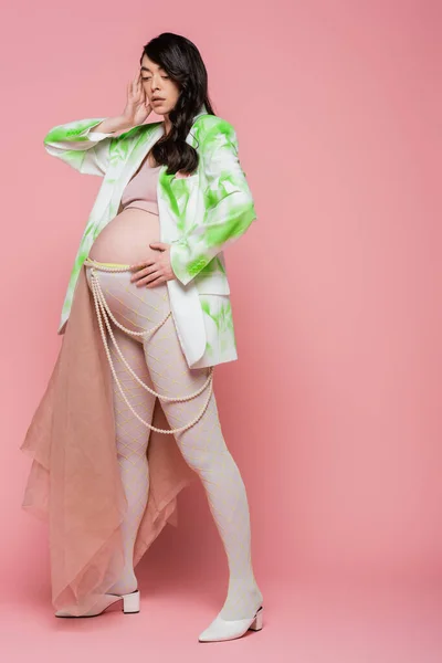 Полная длина модной будущей матери в зеленом и белом блейзере, топ, ремень из бусин и колготки с шифоновой тканью, держа руку возле лица на розовом фоне, концепция моды на беременность — стоковое фото