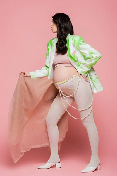 Longitud completa de la futura madre en blazer verde y blanco, top de la cosecha, cinturón de cuentas y leggings posando con tela de gasa beige sobre fondo rosa, concepto de embarazo de moda - foto de stock