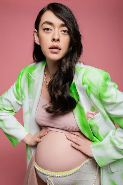 Atractiva mujer embarazada con el pelo ondulado morena, en blazer verde y blanco, top de la cosecha y cinturón de cuentas, posando con las manos en la barriga y mirando a la cámara aislada en rosa, concepto de moda de maternidad - foto de stock